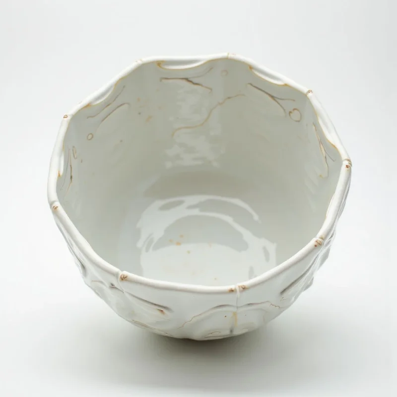 A roughly polygonal, off-white ceramic bowl sits against a pale background. The exterior of the bowl is textured with raised, organic shapes resembling branches or roots and has subtle golden markings. Its interior is smooth and glossy, reflecting light in swirling patterns.