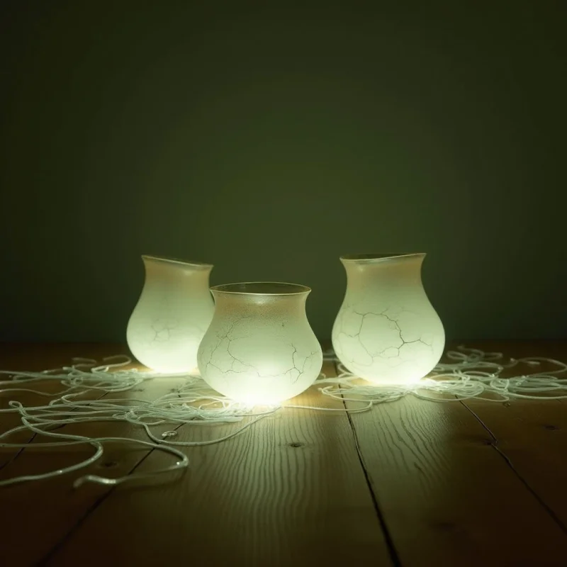Three translucent, bulbous white vessels sit in a row atop a warm-toned wooden surface against a dark, muted green background. The vessels feature delicate, vein-like crackle textures and emit a soft internal glow that illuminates the wood grain below. Tangled thin white wires sprawl across the foreground, connecting the luminous shapes within a shallow spatial plane.